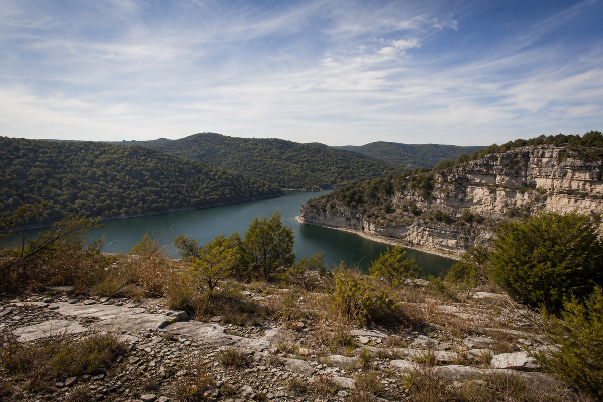 Wooded Ozark hills above clear green lake water with a pale limestone and dolomite bluff face and an open rocky glade in the foreground